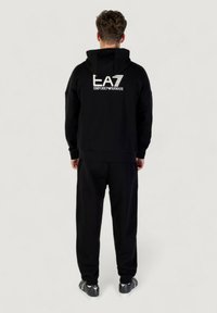 Schwarzer Kapuzenpullover und passende Hose, aus weichem Material, mit "EA7 Emporio Armani"-Logo auf dem Rücken. Die Sneaker haben graue Akzente.