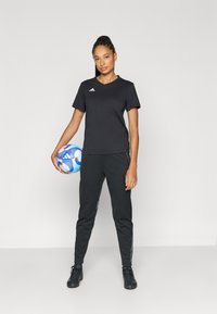 Tricou sportiv negru și pantaloni, cu accente gri pe lateral, purtați de o persoană care ține o minge de fotbal alb-albastră. Încălțămintea neagră completează ținuta.