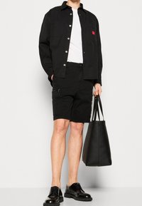 Man i svart knäppt jacka, vit T-shirt, svarta shorts, svarta läderskor och håller en stor svart tygväska.