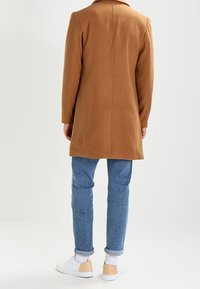 Brun kappa i ullblandning med en skräddarsydd silhuett, utrustad med en notcht krage och en rak rygg. Kombinerad med ljusblå jeans och vita sneakers.