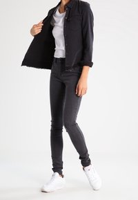 Černá džínsová bunda s roztrhanými lemy, nošená přes bílé tričko. Šedé úzké džíny a bílé tenisky dotvářejí celý outfit.