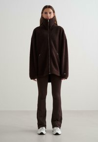 Bruine fleecejas met hoge kraag, ritssluiting en zijzakken. Losse pasvorm, gestructureerde stof, gecombineerd met donkerbruine broek en witte sneakers.