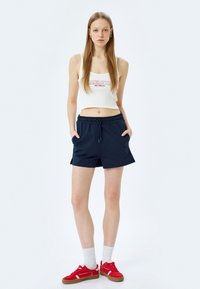 Weißes, geripptes Crop-Top mit dem Text "MANHATTAN", kombiniert mit navyblauen Shorts mit elastischem Bund. Getragen mit roten Sneakers und weißen Crew-Socken.