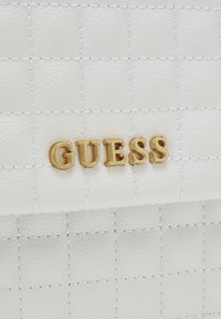 Borsa in pelle sintetica bianca trapuntata con logo dorato "GUESS", accentuata da cuciture bianche e texture a motivi quadrati.