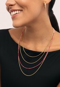 Persona sonriente con pendiente de aro dorado y collares de cuentas doradas y rosas en capas, sobre una camiseta negra, cabello oscuro recogido hacia atrás.