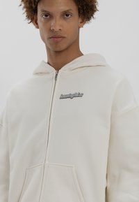 Felpa bianca con zip realizzata in tessuto morbido, dotata di una grande tasca frontale e un logo argentato sul lato sinistro del petto, con cappuccio regolabile.