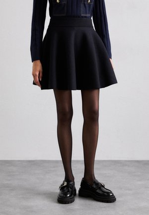 Falda negra acampanada con una cinturilla ancha, combinada con medias negras opacas y mocasines negros brillantes con un detalle de hebilla metálica.