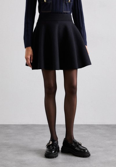 Jupe noire évasée avec une large ceinture, associée à des collants noirs opaques et des mocassins noirs brillants avec un détail de boucle en métal.