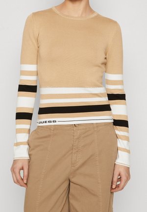 Pull beige à manches longues avec des rayures horizontales blanches et noires et le texte "GUESS" près de l'ourlet, porté avec un pantalon beige.
