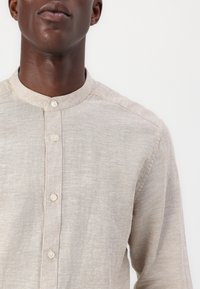 Camisa beige de manga larga con cuello alzado, hecha de una tela suave y texturada; presenta una tapeta frontal con botones de color claro.