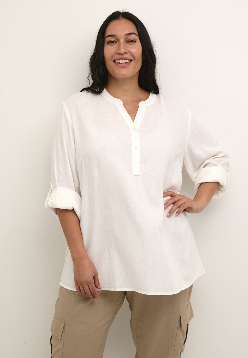 Chemise en lin blanc avec un col mandarin, trois boutons et des manches retroussées, associée à un pantalon cargo beige clair. Texture lisse et coupe ample.
