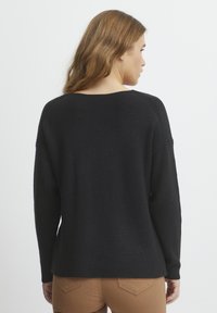 Pull noir à côtes avec une coupe ample et un grand col, doté de manches longues et de coutures d'épaule abaissées, porté sur un pantalon marron clair.