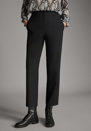 Frau trägt schwarze, gerade geschnittene Hose, schwarze Schnürstiefel bis zum Knöchel und eine langärmlige Bluse mit Schlangenmuster, die Hände in den Taschen.