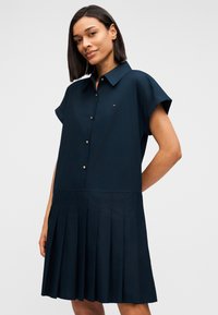 Φόρεμα πουκαμίσα σε ναυτικό μπλε χρώμα με κοντά μανίκια, κουμπιά στο μπροστινό μέρος και πλισέ φούστα. Διαθέτει μικρό λογότυπο στο στήθος και υφασμάτινη υφή.
