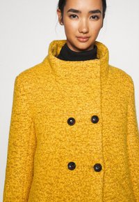 Cappotto giallo in lana con finitura testurizzata, colletto alto, design doppiopetto e quattro bottoni neri.
