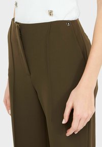 Pantaloni verde oliva su misura con una texture liscia, dotati di chiusura con zip, tasche laterali e dettagli metallici in vita.