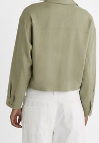 Jasnozielona, długorękawowa koszula z teksturowanej tkaniny, z mankietem na guzik i lekko skróconym tyłem; zestawiona z białymi spodniami.