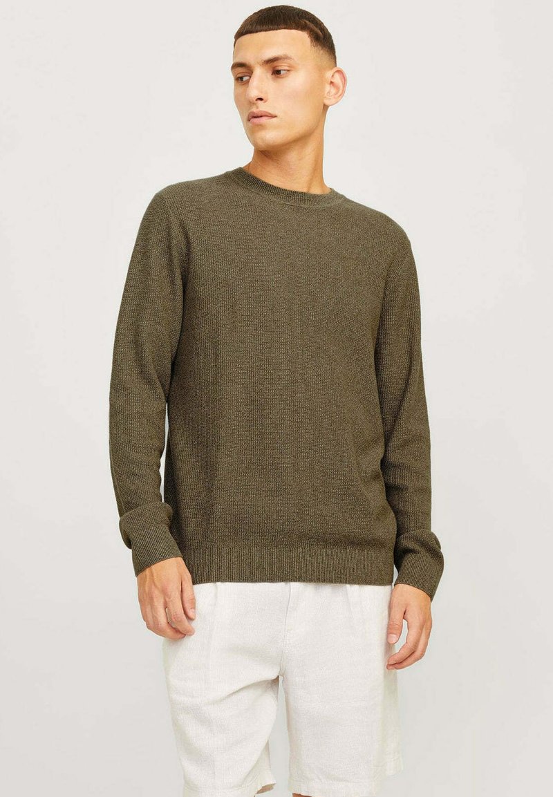 Pull en maille vert olive avec un col rond, des poignets et un ourlet côtelés. Texture douce, design ajusté, porté avec un short de couleur claire.