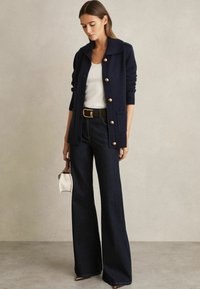 Cardigan tricotat bleumarin cu butoane mari, asociat cu un top alb, blugi largi negri și o geantă mică albă.