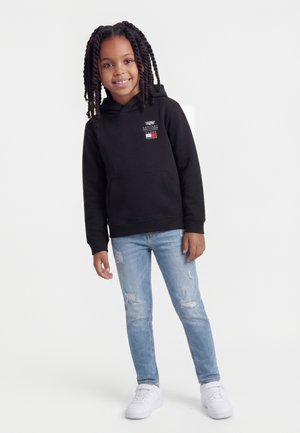 Enfant souriant aux longues tresses portant un sweat à capuche noir, un jean clair déchiré et des baskets blanches, debout devant un fond blanc uni.