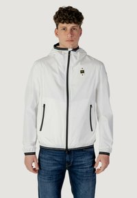 Giacca leggera bianca con zip sul davanti, cappuccio e dettagli neri su tasche e polsini. Presenta un logo sul petto.