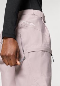 Houdini kültéri nadrág világos rózsaszínben, oldalán cipzáras zsebbel, sima textúrával és varrási részletekkel, valamint logóval a derékrészen.