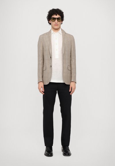 Hellbrauner kariert Blazer über einem weißen Leinenhemd, kombiniert mit schwarzen Hosen und schwarzen Schuhen. Model trägt dunkle Sonnenbrillen.