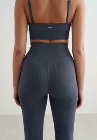 Sivý rebrovaný crop top a legíny s vysokým pásom s vertikálnymi pruhmi. Obsahuje nastaviteľné ramienka a malú logo dosku na chrbte.