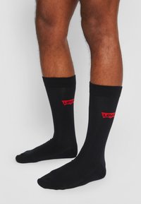 Sorte bomuldsstrømper med ribbede kanter, der har et rødt "Levi's" logo på fronten. Simpelt design, længde til midten af læggen, glat tekstur.