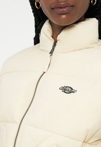 Giacca puffer crema con colletto alto, dotata di zip frontale e logo "Dickies" nero sul petto. Tessuto trapuntato con tasche laterali.