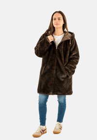 Cappotto in pelliccia sintetica marrone con cappuccio, dotato di zip frontale e tasche laterali. Indossato sopra una maglietta bianca e jeans blu, abbinato a scarpe da ginnastica beige.