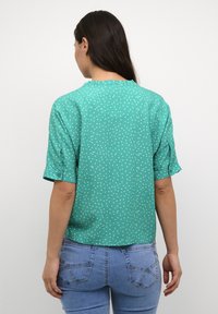 Blusa turchese con pois bianchi, maniche corte e dettaglio nella scollatura arricciato. Realizzata in tessuto leggero, presenta maniche arrotolate e una vestibilità comoda.