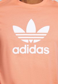 Ljusorange t-shirt med en stor vit Adidas-logotyp med en trebladig design och tre horisontella ränder under varumärkets namn.