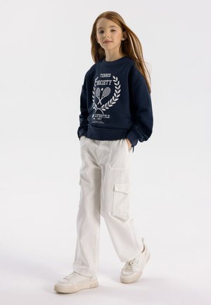 Felpa navy con stampa bianca che presenta racchette da tennis, abbinata a pantaloni cargo grigio chiaro e sneakers bianche con suole testurizzate.