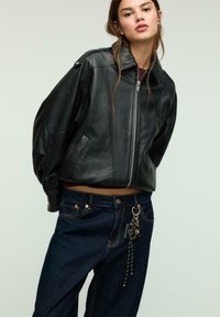 Giacca bomber in pelle nera con chiusura a zip, spalle larghe e tasche laterali. Abbinata a jeans scuri in denim con un accento di portachiavi metallico.