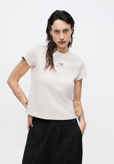 Vaaleanpunainen puuvillainen t-paita, jossa on lyhyet hihat ja pyöreä kaula-aukko sekä pieni graafinen kuvio rintakehässä. Yhdistetty mustiin housuihin.