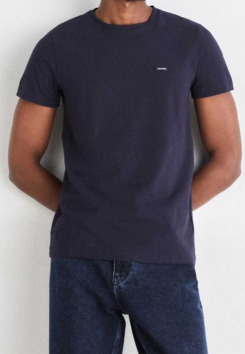 Homme portant un t-shirt Calvin Klein uni bleu marine foncé, non rentré, sur un jean bleu foncé, mains derrière le dos, debout devant un fond blanc.