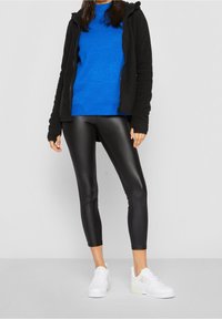 Sudadera negra con cremallera sobre un suéter de punto azul, combinada con leggings brillantes negros y zapatillas blancas con acentos texturizados.