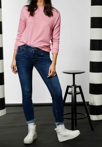 Jasnoróżowy sweter z dzianiny, dopasowane ciemnoniebieskie dżinsy oraz białe sneakersy za kostkę. Tło zdobią pionowe paski w czerni i bieli.