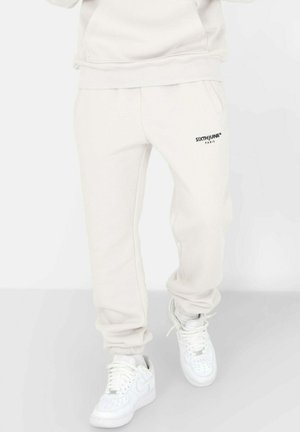 Helle beige Jogginghose aus weichem Stoff, mit Seitentaschen und einem kleinen schwarzen Logo am linken Bein. Kombiniert mit weißen Sneakers.