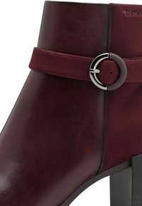 Bottine en cuir bordeaux avec une large sangle ornée d'une boucle en argent circulaire, texture lisse et un talon épais noir.