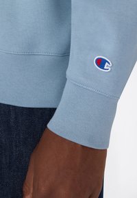 Manga de sudadera azul clara con parche del logo de Champion cerca de la muñeca, en una persona que lleva jeans azul oscuro.