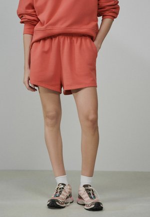 Person trägt korallfarbene Shorts und ein passendes Sweatshirt, weiße Socken und hellrosa Laufschuhe, steht auf einem grauen Boden.