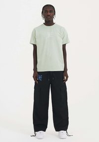 Camiseta de algodón verde claro con un diseño gráfico blanco, combinada con pantalones cargo negros sueltos que tienen un logotipo azul y múltiples bolsillos. Zapatillas blancas.