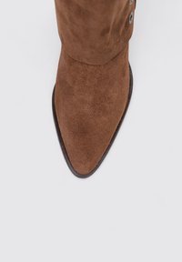 Botín de ante marrón hasta el tobillo con punta afilada, textura suave y botones plateados decorativos en el costado. Suela plana y oscura visible.