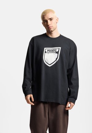 Młody mężczyzna z krótkimi blond włosami, ubrany w czarną koszulę z długim rękawem z białym logo tarczy "PROSTO" oraz brązowe spodnie, stojący na białym tle.