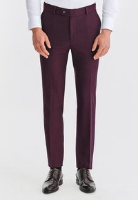 Mężczyzna noszący ciemnofioletowe dopasowane spodnie garniturowe, białą koszulę oraz czarne skórzane półbuty sznurowane, stoi na tle jednolitego tła.