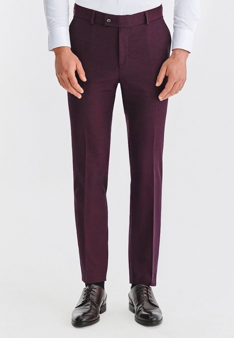 Mężczyzna noszący ciemnofioletowe dopasowane spodnie garniturowe, białą koszulę oraz czarne skórzane półbuty sznurowane, stoi na tle jednolitego tła.