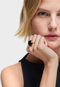 Anillo de oro con una gran piedra negra facetada, montado en una banda pulida con cuatro acentos esféricos cerca de la piedra. Mano posicionada en la mejilla.