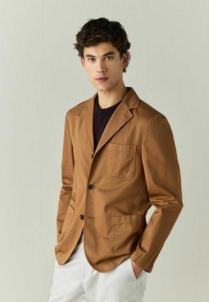 Ung mand med mørkt krøllet hår iført en brun blazer over en mørk sweater og hvide bukser, stående mod en ensfarvet lys baggrund.